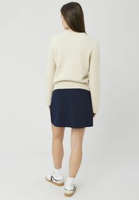 Pull beige clair, texture tricotée, col rond ; associé à une jupe bleu marine, tissu lisse ; des baskets blanches et des chaussettes complètent la tenue.