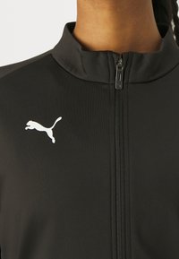 Svart zip-up jakke laget av glatt stoff med høy krage og en hvit Puma-logo på venstre bryst.