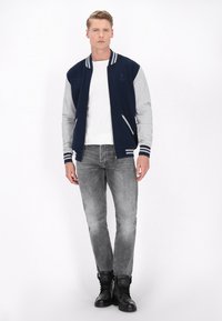 Marineblå og grå varsity jakke med ribbede krave og manchetter, lynlåslommer, båret over en hvid skjorte, parret med grå jeans og sorte støvler.