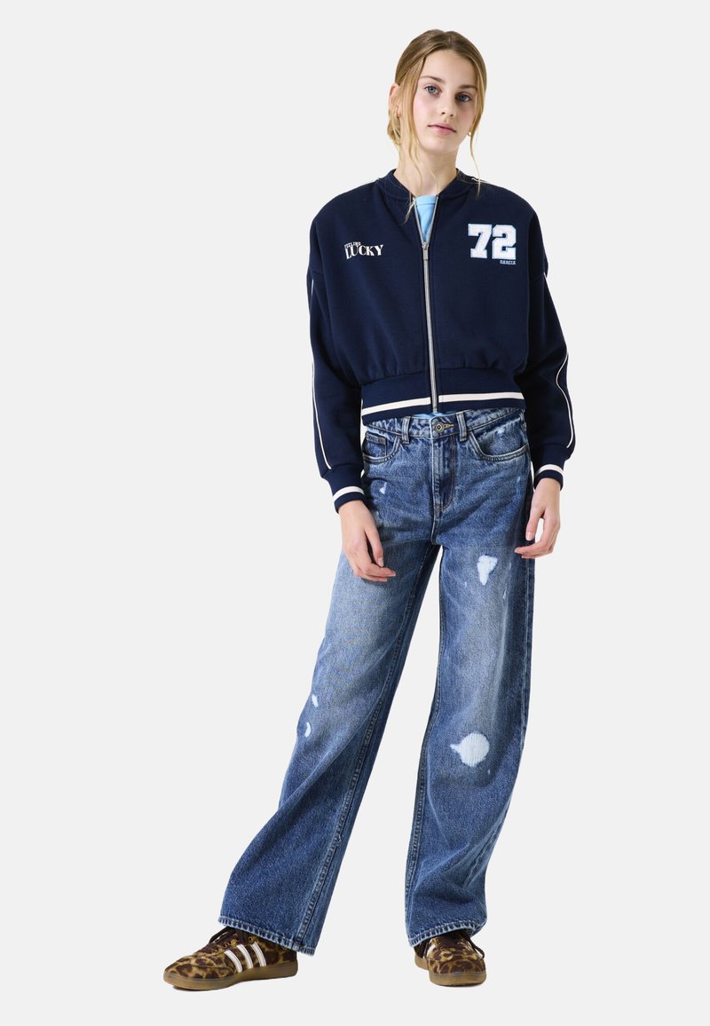 Veste bomber coupée bleu marine avec des rayures blanches et du texte ; jean bleu à large jambe déchiré ; baskets brunes avec des rayures et des accents de motif animal.