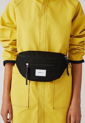 Personne portant une veste jaune aux manches retroussées et un sac banane noir Sandqvist attaché autour de la taille.