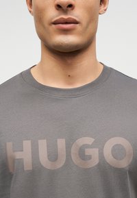 Κοντινό πλάνο ενός ατόμου που φοράει γκρι μπλουζάκι με στρογγυλή λαιμόκοψη και το όνομα της μάρκας "HUGO" τυπωμένο στο στήθος.