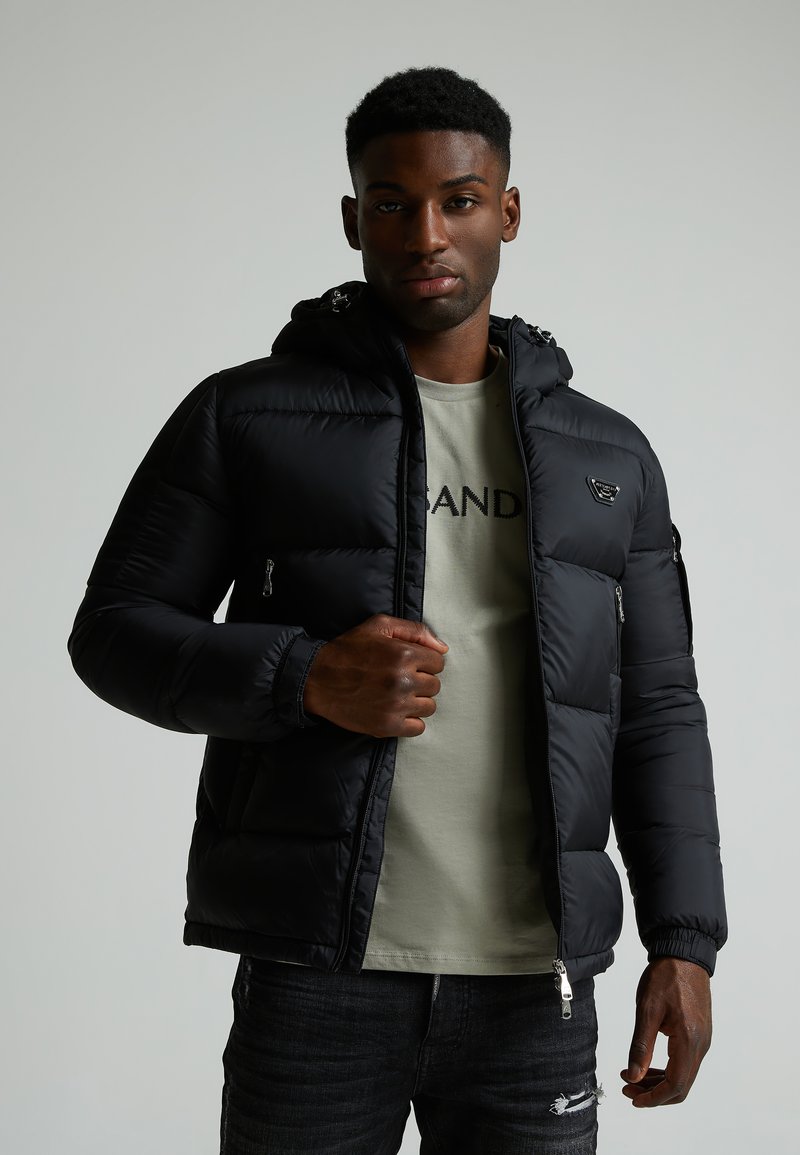 Schwarze kapuzenlose Jacke aus Puffermaterial mit Reißverschluss-Taschen und elastischen Bündchen, getragen über einem hellgrünen Hemd, kombiniert mit dunklen Jeans.