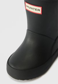 Bota de borracha preta com uma sola texturizada preta, bico arredondado e um rótulo vermelho e branco "HUNTER" na lateral. Acabamento suave no geral.