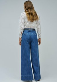 Mujer con cabello ondulado vistiendo una blusa de encaje blanco y jeans de pierna ancha de talle alto en azul con un cinturón de cadena, de pie contra un fondo liso.