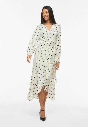 Kvinne som går i en hvit omslagskjole med svarte prikker og lange ermer, iført svarte hæler med ankelstropp, smilende svakt til siden.