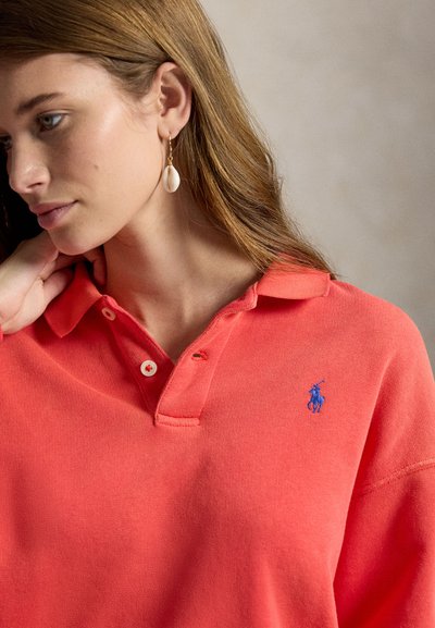 Mujer con cabello largo y castaño, vestida con una camiseta polo coral con un logo azul bordado y un pendiente de aro dorado con un colgante de concha blanca, mirando hacia abajo.