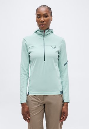 Kobieta ubrana w jasnozielony, długorękawowy pulower z kapturem i zamkiem z przodu oraz beżowe spodnie, stojąca na jednolitym tle.