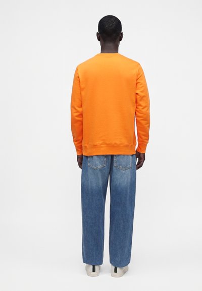 Homme de dos portant une chemise orange vif à manches longues, un jean bleu ample et des chaussures blanches sur un fond uni clair.