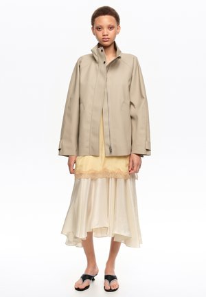 Persona vestida con chaqueta beige con cremallera sobre un vestido de encaje crema y amarillo con volantes, y sandalias de tacón negras abiertas, de pie contra un fondo blanco.