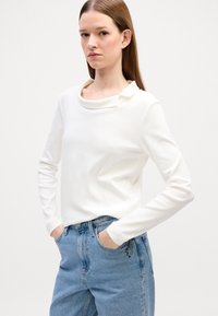 Witte langslavige top met een colkraag en subtiele plooidetails, gecombineerd met hooggewaist blauwe denimjeans. Over het geheel een soepele textuur.