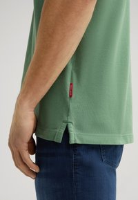 Maglietta di cotone verde con finitura testurizzata, fessure laterali all'orlo e un'etichetta di marca rossa sul lato sinistro inferiore, indossata con jeans blu.