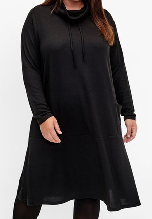 Femme portant une robe noire ample à manches longues avec un col bénitier et une poche avant, debout devant un fond blanc.