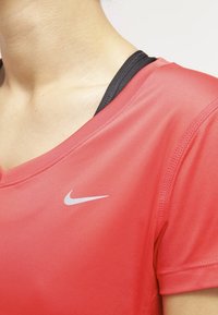 Nike urheilu paita kirkkaan punaisena, jossa on V-kaula-aukko, lyhyet hihat ja pieni hopeinen logo vasemmalla puolella. Tasainen, kevyt kangas.
