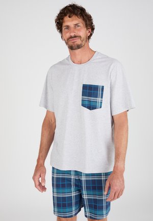 Man draagt een lichtgrijs T-shirt met een blauw geruit zakje en bijpassende blauw geruite korte broek, staand tegen een effen achtergrond.