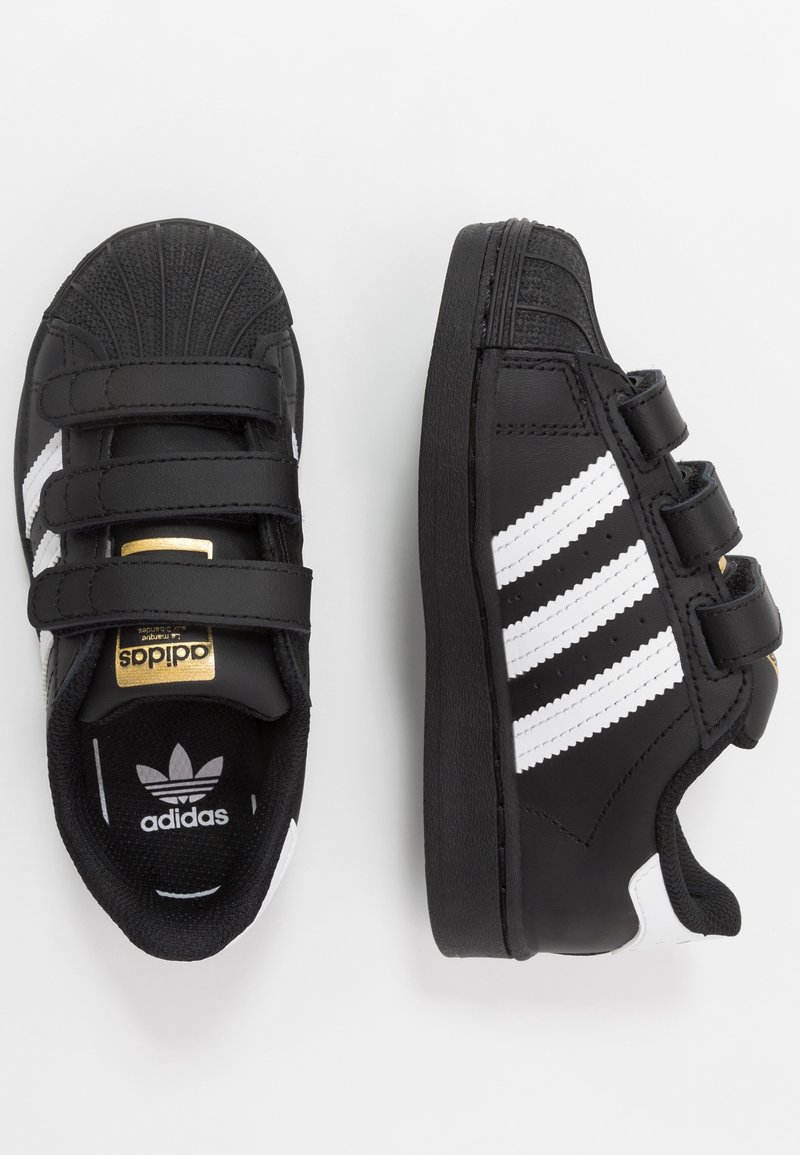 Baskets Adidas noires avec des bandes blanches, trois bandes Velcro, et un logo doré sur la languette, présentées en vues de dessus et de côté.