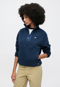 Marineblå fleecegenser med halvglidelås, med teksturert overflate og en liten logo på brystet, kombinert med khakifargede bukser.