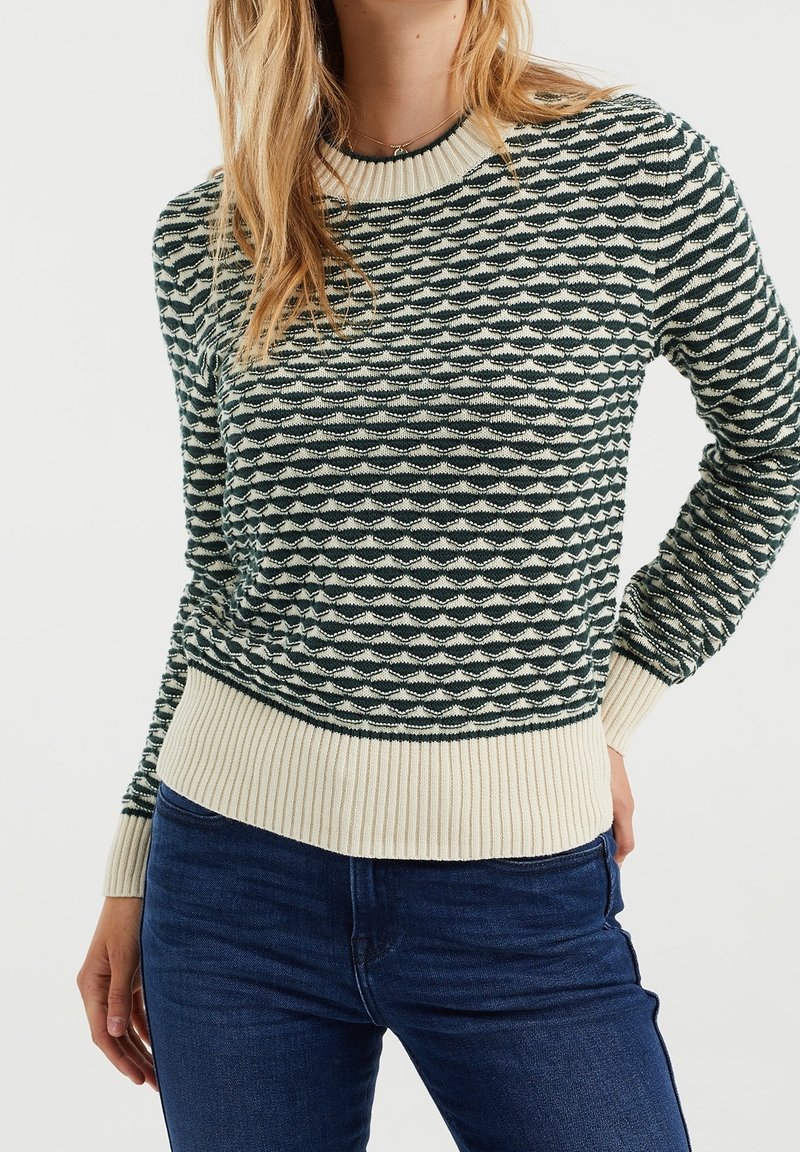 Femme portant un pull en maille à manches longues avec un motif ondulé noir et blanc, des poignets et un ourlet côtelés, associé à un jean bleu foncé.
