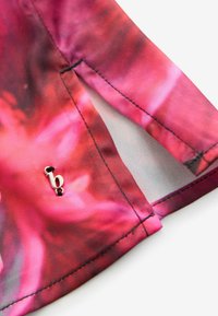 Pink mønstret stof hjørne med glat tekstur, der har et lille metallisk logo med sorte accenter nær sømmen.