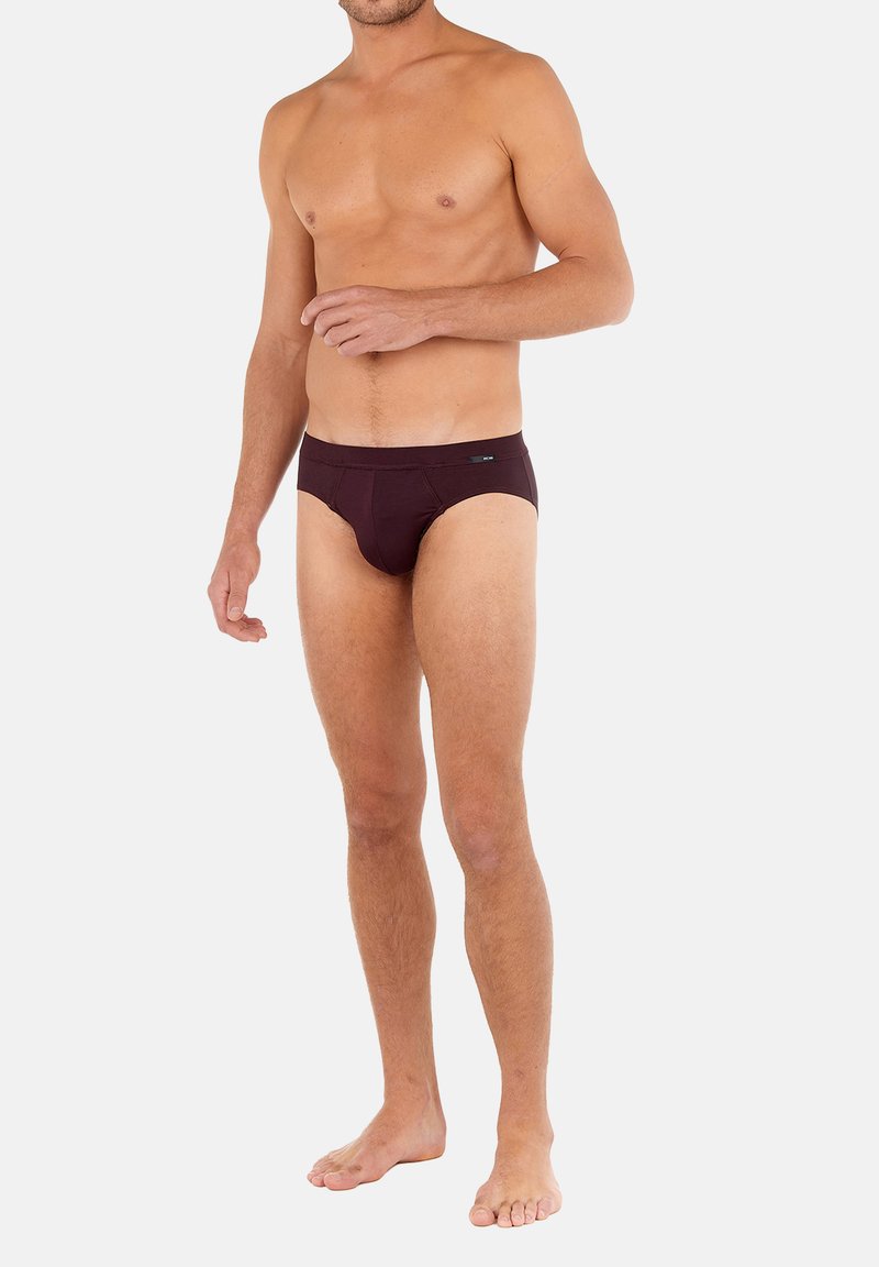 Oben ohne männliches Model, das barfuß steht und dunkellilafarbene Unterwäsche trägt, vor einem schlichten weißen Hintergrund.