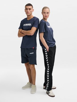 Man in marineblauwe Hummel shorts en T-shirt met gekruiste armen staat rug aan rug met vrouw in marineblauwe Hummel T-shirt en zwarte broek met zij-chevronstrepen.