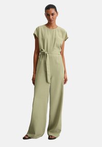 Jumpsuit verde chiaro con una vestibilità morbida, maniche corte, una tasca frontale e una cintura coordinata in vita. Tessuto leggero con una consistenza liscia.