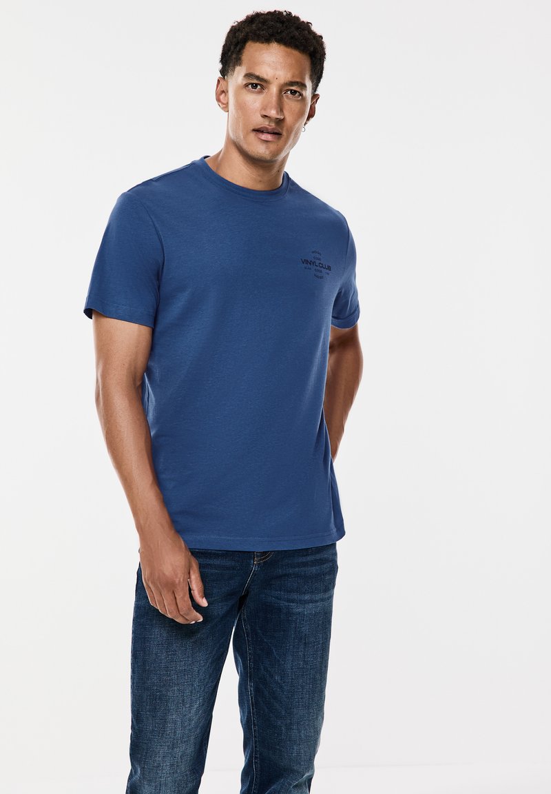 Sininen puuvillainen t-paita, jossa on lyhyet hihat ja pyöreä kaula-aukko, jossa rintaosassa on mustalla painettuna teksti "VINYL CLUB". Asusteena t-paidan kanssa tummat farkut.