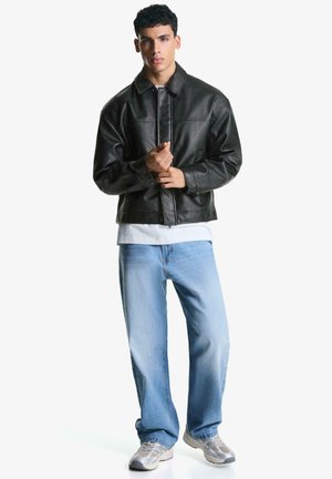 Schwarze Lederjacke mit Kragen, ausgestattet mit einem Reißverschluss. Kombiniert mit hellblauen Jeans und grauen Sportschuhen.