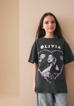 Ung person med langt brunt hår iført en oversized sort T-shirt med "OLIVIA" og et hjerteformet motiv af en kvinde og roser, kombineret med lyse jeans.