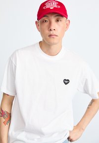 White cotton t-shirt with short sleeves, round neckline, and black heart-shaped logo on the left chest. Worn with a red cap.