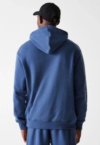 Sweat à capuche en coton bleu avec une grande capuche, des poignets et un ourlet côtelés, présentant une texture lisse et une coupe décontractée. Porté avec un pantalon assorti bleu.