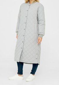 Donna che indossa un lungo cappotto grigio chiaro imbottito con bottoni a pressione, jeans skinny scuri e sneakers bianche, che si trova contro uno sfondo bianco.