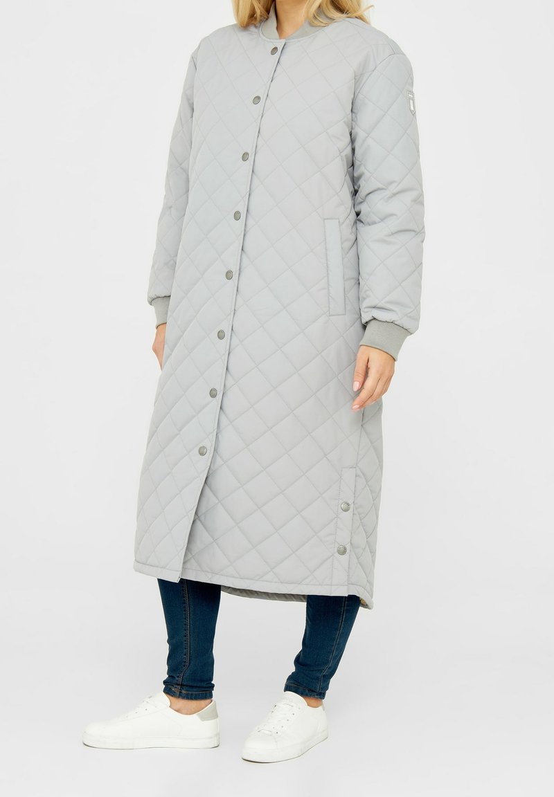 Donna che indossa un lungo cappotto grigio chiaro imbottito con bottoni a pressione, jeans skinny scuri e sneakers bianche, che si trova contro uno sfondo bianco.
