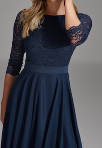 Kvinna i en marinblå klänning med spetstopp och trekvartslånga ärmar, samt en slät, flödande kjol.