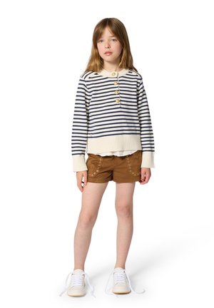 Dziewczyna stoi ubrana w kremowy sweter w granatowe paski z złotymi guzikami, brązowe szorty z oczkami oraz białe trampki z rozwiązanymi sznurówkami.
