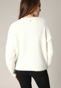 Witte, losse gebreide trui met een zachte textuur, ronde hals en verlaagde schouders. Kleine hartdetail aan de achterkant van de halslijn.
