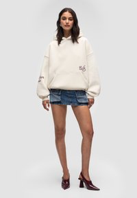 Kremowa hoodie z przednią kieszenią i haftowanym "W" na prawym rękawie, w połączeniu z dżinsową mini spódnicą i burgundowymi butami na obcasie.