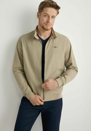 Mężczyzna z jasnobrązowymi włosami, ubrany w beżową kurtkę na zamek błyskawiczny na czarnej koszulce i ciemnoniebieskich spodniach, stojący na białym tle.