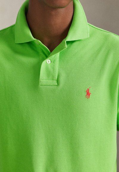Grønn poloskjorte i strukturert bomull med krage og knappestolpe. Et rødt brodert logo er på venstre bryst.