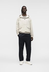 Ελαφρύ μπεζ μπουφάν με κουκούλα, εμπρόσθιο φερμουάρ και τσέπες με πατς, συνδυασμένο με σκούρο παντελόνι και λευκά αθλητικά παπούτσια. Ομαλή υφή σε όλο το ύφασμα.