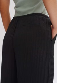Black pleated pants with an elastic waistband, featuring a back pocket detail and side pockets, made from textured fabric.