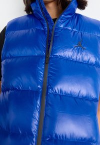Blå pufferväst med hög krage, glänsande nylonmaterial, dragkedja fram och svarts logoaccent på vänster bröst. Texturerad vaddering synlig.