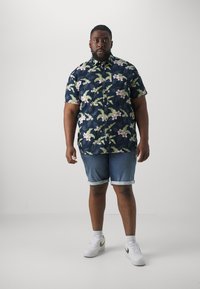 Lyhytha sleevesi, tummansininen kukallinen paita, jossa on vihreitä lehtiä ja vaaleanpunaisia kukkia, yhdistettynä vaaleansinisiin denim-shortsisiin ja valkoisiin lenkkikenkiin, joissa on kultaiset yksityiskohdat.