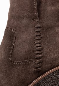 Brun mocka stövel med en texturerad yta, sidostygn och en dekorativ skärsöm. Gummisula med ett mönstrat grepp.