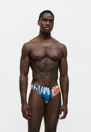 Homme musclé debout, face à l’avant, portant un slip de bain coloré avec un motif de texte abstrait bleu, blanc et orange sur fond blanc.