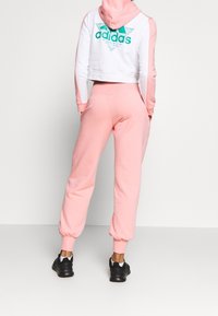 Lyhennetty valkoinen ja vaaleanpunainen huppari, jossa on turkoosi Adidas-logo, yhdistettynä matching-vaaleanpunaisiin verkkareihin, kerroksellisiin hihoihin ja mustiin lenkkareihin.