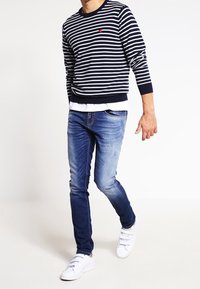 Randig marinblå och vit sweatshirt med rundad hals, lager på lager över en vit t-shirt; kombinerad med ljusblå jeans och vita sneakers.