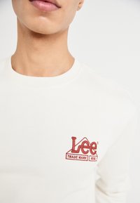 Felpa crema in misto cotone, con logo "Lee" ricamato in rosso. Collo rotondo e polsini a coste. Design semplice e pulito.