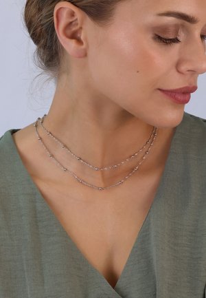 Silberne mehrschichtige Halskette mit strukturierten Gliedern und kleinen runden Akzenten, präsentiert vor einem sanften, gedämpften grünen Kleidungsstück.