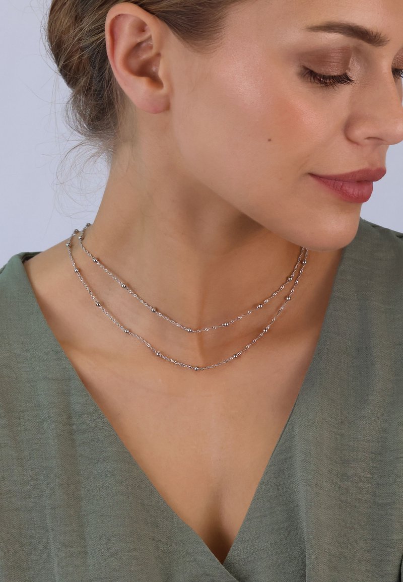 Silberne mehrschichtige Halskette mit strukturierten Gliedern und kleinen runden Akzenten, präsentiert vor einem sanften, gedämpften grünen Kleidungsstück.